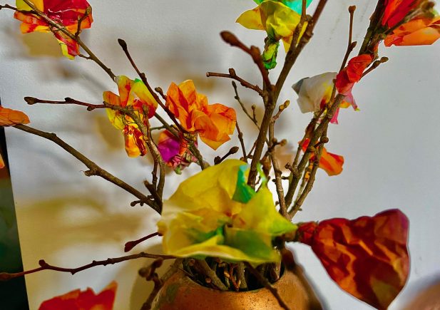 Die in einer Vase arrangierten Zweige sind mit bunten Seidenpapierblumen in Orange-, Gelb-, Grün- und Rottönen geschmückt, die vor einem unifarbenen Hintergrund ein fröhliches Bild ergeben.