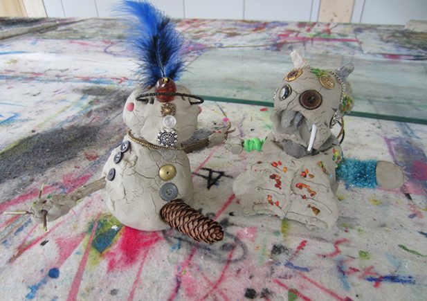Zwei kleine Tonfiguren, die mit Knöpfen, Tannenzapfen und anderen Bastelmaterialien verziert sind, sitzen auf einer bunten, mit Farbe bespritzten Oberfläche. Eine hat eine blaue Feder an der Spitze und die andere scheint Stoßzähne und verschiedene Knöpfe zu haben.