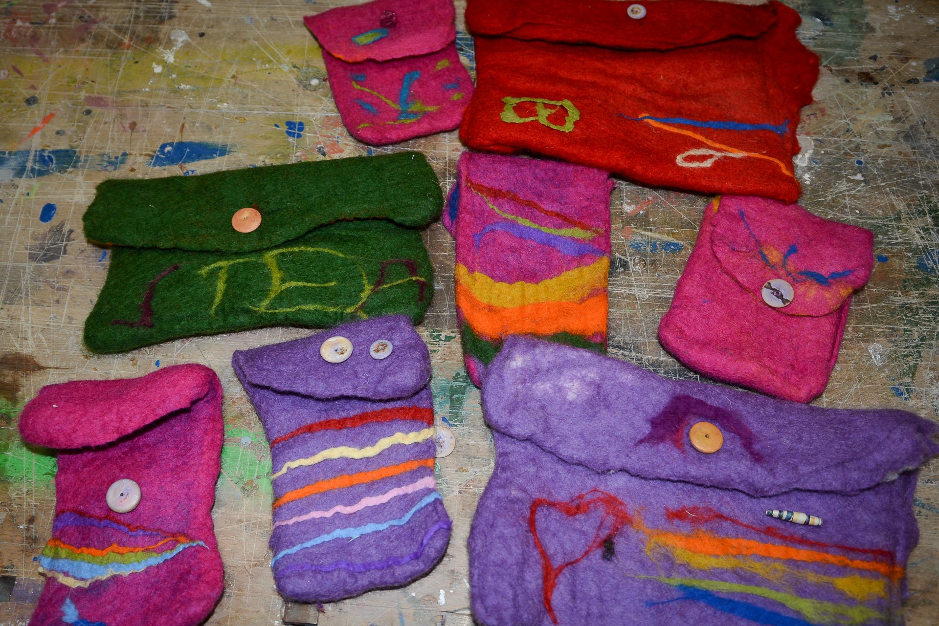 Mehrere bunte, handgefertigte Filztaschen mit Knöpfen und verschiedenen abstrakten Mustern liegen auf einem mit Farbe bespritzten Tisch. Die Taschen sind hauptsächlich lila, rosa, rot und grün mit bunten Akzenten.