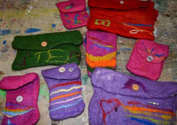 Mehrere bunte, handgefertigte Filztaschen mit Knöpfen und verschiedenen abstrakten Mustern liegen auf einem mit Farbe bespritzten Tisch. Die Taschen sind hauptsächlich lila, rosa, rot und grün mit bunten Akzenten.