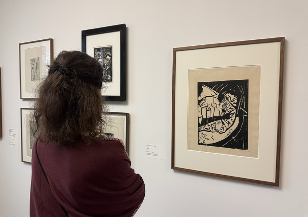 Eine Person mit lockigem Haar und kastanienbraunem Schal steht vor gerahmten Kunstwerken in einer Galerie und betrachtet einen Schwarz-Weiß-Druck, der eine klavierspielende Person zeigt. Andere gerahmte Werke hängen an der weißen Wand daneben.