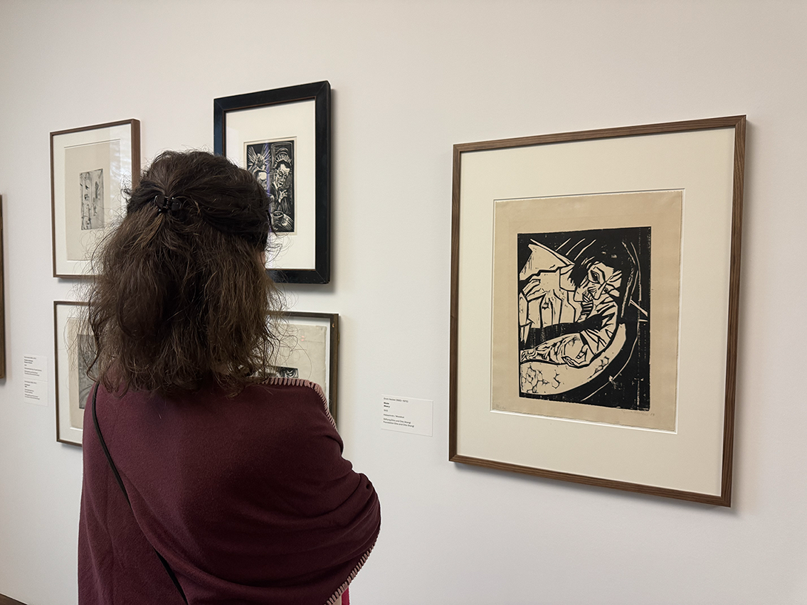 Eine Person mit lockigem Haar und kastanienbraunem Schal steht vor gerahmten Kunstwerken in einer Galerie und betrachtet einen Schwarz-Weiß-Druck, der eine klavierspielende Person zeigt. Andere gerahmte Werke hängen an der weißen Wand daneben.