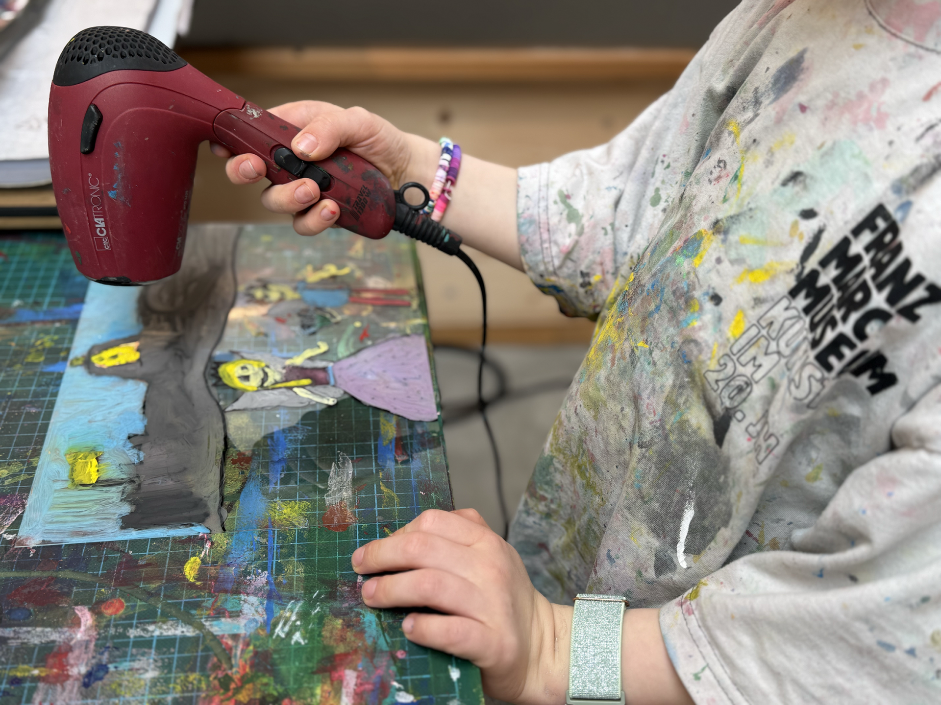 Ein Kind verwendet einen roten Föhn, um ein gemaltes Kunstwerk auf einem unordentlichen Kunsttisch zu trocknen. Das Kind trägt ein mit Farbe bespritztes Hemd und ein glitzerndes Armband, im Hintergrund ist buntes Kunstzubehör zu sehen.