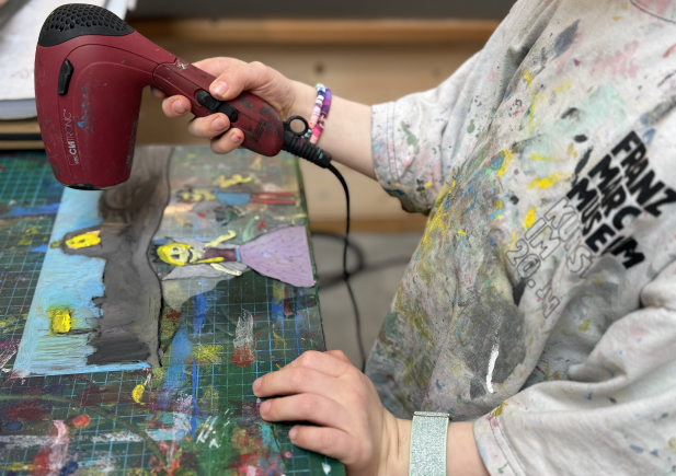 Ein Kind verwendet einen roten Föhn, um ein gemaltes Kunstwerk auf einem unordentlichen Kunsttisch zu trocknen. Das Kind trägt ein mit Farbe bespritztes Hemd und ein glitzerndes Armband, im Hintergrund ist buntes Kunstzubehör zu sehen.