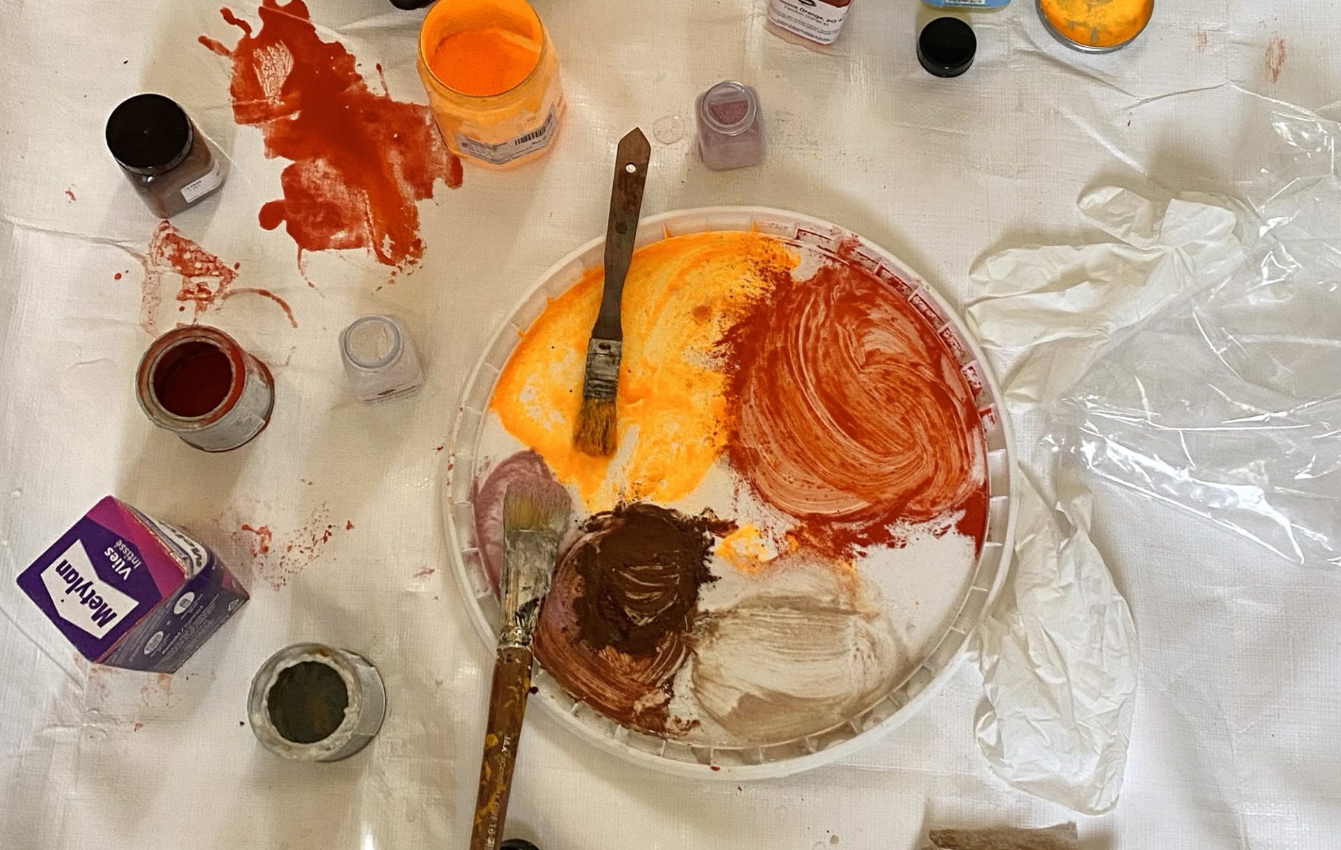 Ein unordentlicher Arbeitsbereich eines Künstlers mit Farbtöpfen, offenen Deckeln, einer mit orangefarbener, roter, brauner und weißer Farbe verschmierten Plastikpalette, zwei Pinseln, Spritzern auf einem weißen Tuch und einem zerknitterten Plastikhandschuh.