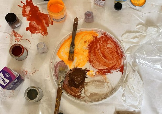 Ein unordentlicher Arbeitsbereich eines Künstlers mit Farbtöpfen, offenen Deckeln, einer mit orangefarbener, roter, brauner und weißer Farbe verschmierten Plastikpalette, zwei Pinseln, Spritzern auf einem weißen Tuch und einem zerknitterten Plastikhandschuh.