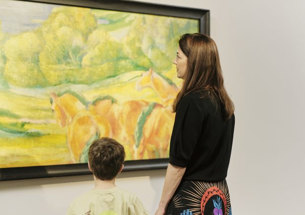 Eine Frau und ein Kind stehen vor einem farbenfrohen Gemälde mit Pferden und Grünpflanzen in einer Kunstgalerie und betrachten das Kunstwerk gemeinsam.