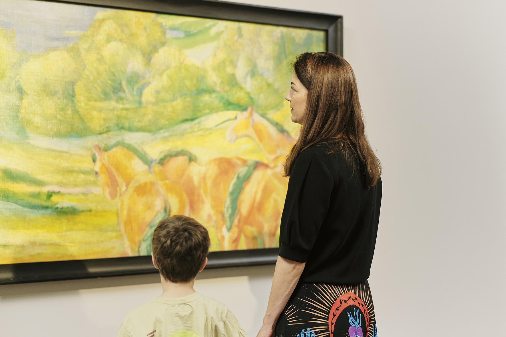 Eine Frau und ein Kind stehen vor einem farbenfrohen Gemälde mit Pferden und Grünpflanzen in einer Kunstgalerie und betrachten das Kunstwerk gemeinsam.