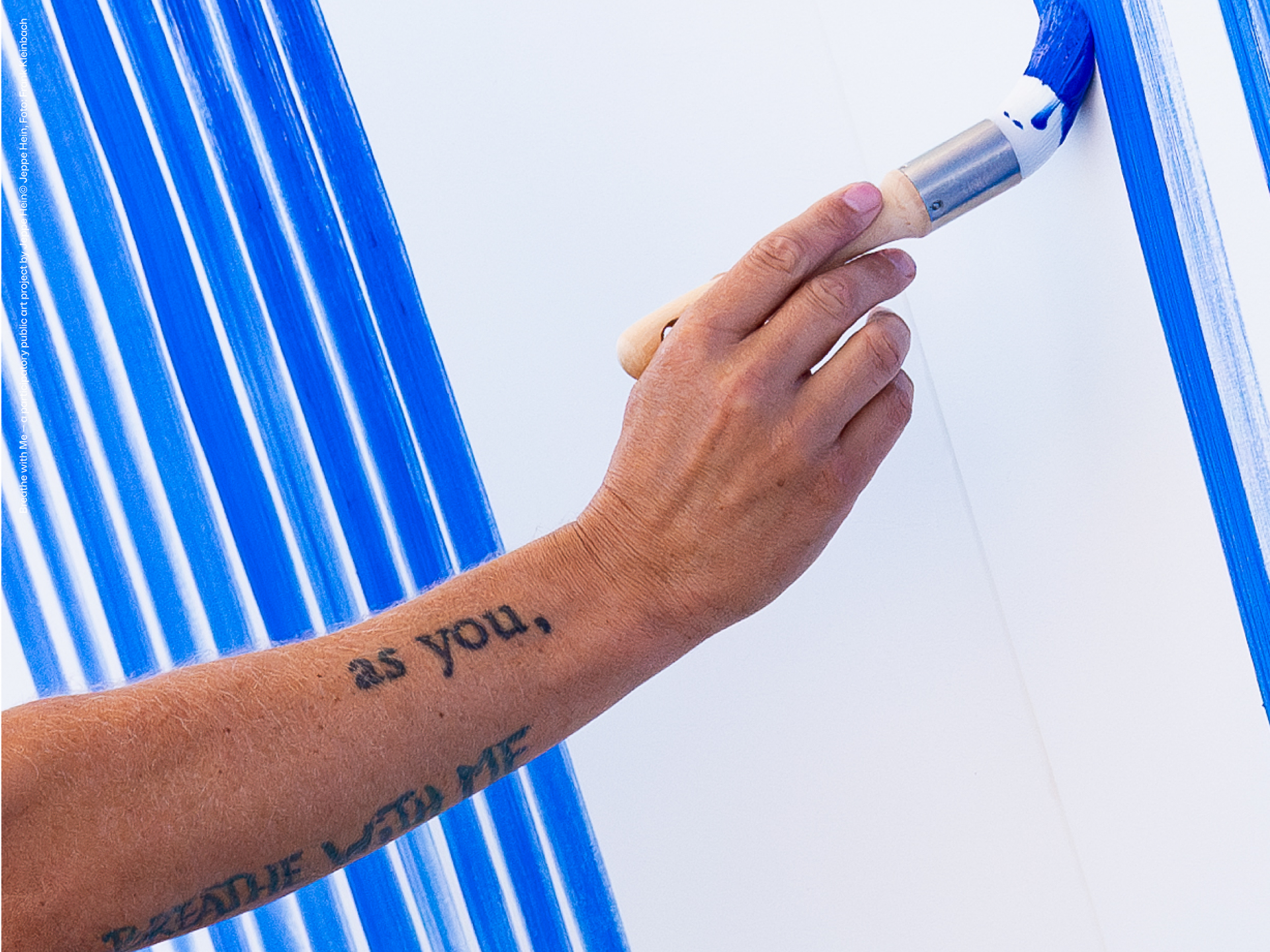 Eine Person mit einem tätowierten Unterarm malt mit einer Farbrolle blaue vertikale Streifen auf eine weiße Wand. Die Tätowierung lautet wie du. und ein Teil des Textes ist unten sichtbar.