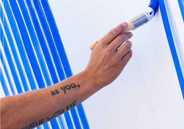 Eine Person mit einem tätowierten Unterarm malt mit einer Farbrolle blaue vertikale Streifen auf eine weiße Wand. Die Tätowierung lautet wie du. und ein Teil des Textes ist unten sichtbar.