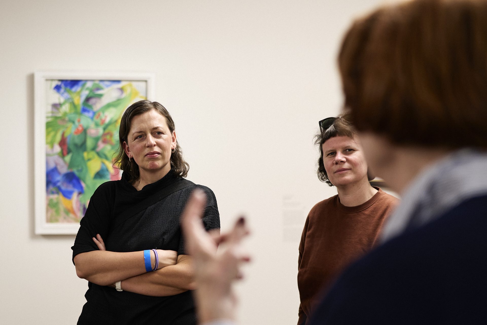 Zwei Frauen stehen aufmerksam in einer Kunstgalerie und hören einer Person zu, die im Vordergrund spricht. Hinter ihnen hängt ein buntes abstraktes Gemälde an der Wand.