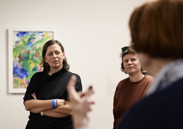 Zwei Frauen stehen aufmerksam in einer Kunstgalerie und hören einer Person zu, die im Vordergrund spricht. Hinter ihnen hängt ein buntes abstraktes Gemälde an der Wand.