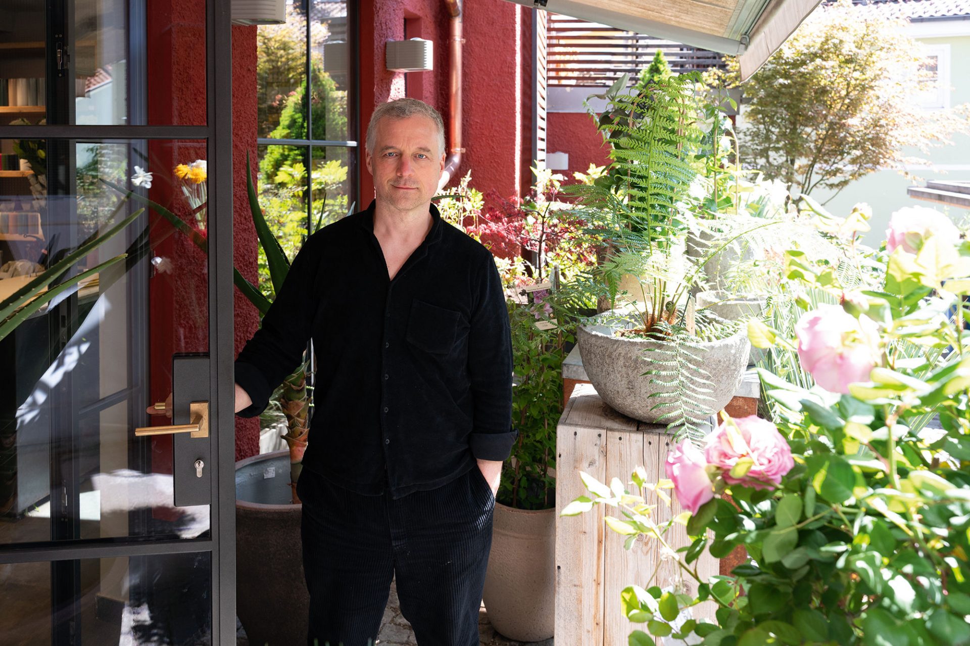 Ein Mann in einem schwarzen Hemd steht an einer offenen Glastür, umgeben von üppigen grünen Pflanzen und rosa Blumen auf einer sonnenbeschienenen Terrasse. Die Szene hat eine entspannte und einladende Atmosphäre.