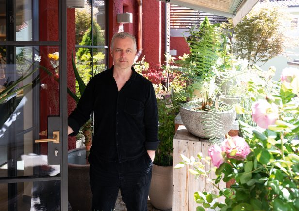 Ein Mann in einem schwarzen Hemd steht an einer offenen Glastür, umgeben von üppigen grünen Pflanzen und rosa Blumen auf einer sonnenbeschienenen Terrasse. Die Szene hat eine entspannte und einladende Atmosphäre.