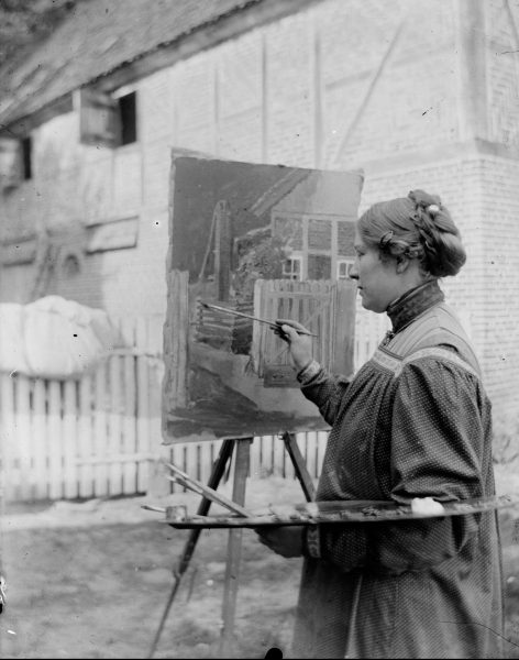 A woman stands outdoors painting a building scene on a canvas set on an easel. She holds a palette and brush, wearing a long dress with an apron. A rustic house and white picket fence are visible in the background.