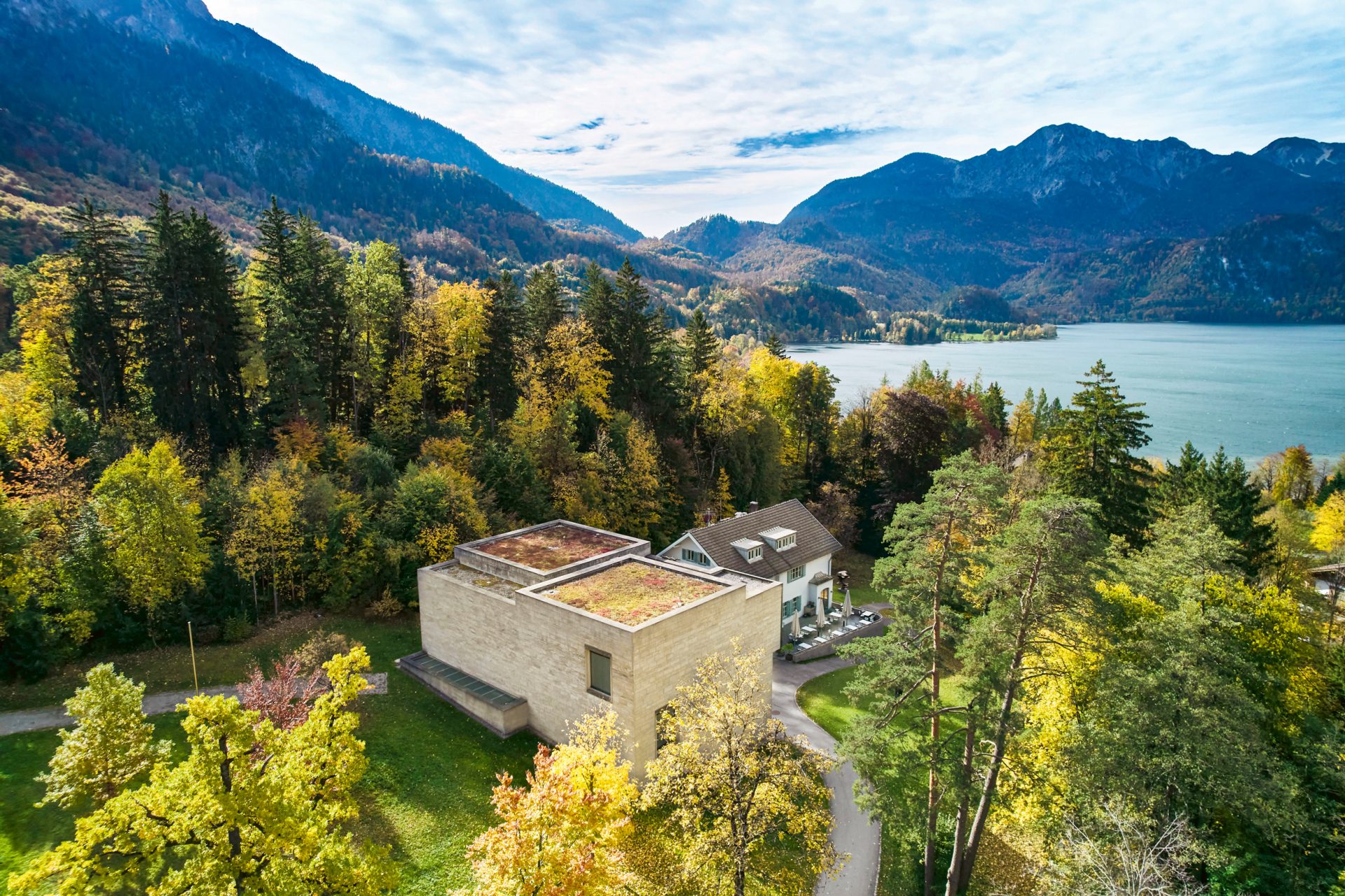 Ein modernes Gebäude mit einem begrünten Dach steht inmitten bunter Herbstbäume in der Nähe eines großen Sees, im Hintergrund sind bewaldete Berge und ein teilweise bewölkter Himmel zu sehen.