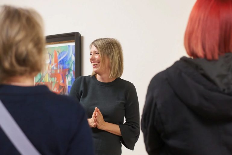 Eine Frau mit blonden Haaren lächelt und faltet die Hände, während sie in einer Kunstgalerie mit zwei Personen im Vordergrund spricht; hinter ihr hängt ein farbenfrohes abstraktes Gemälde an der Wand.