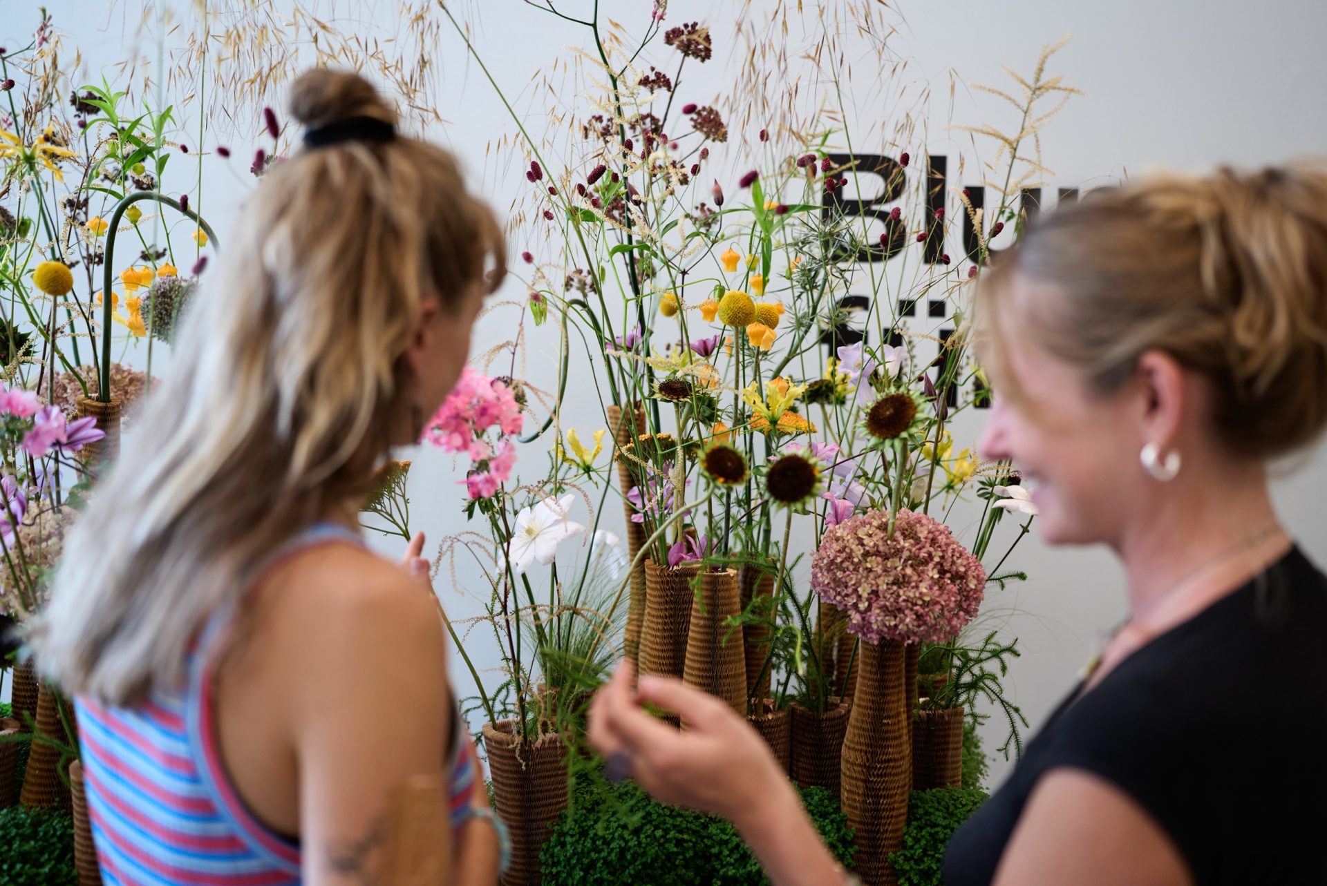 Zwei Frauen stehen neben einem Arrangement aus bunten Blumen in geflochtenen Vasen; eine von ihnen zeigt auf das Arrangement. Der Hintergrund, der von „Zoo und Moderne“ inspiriert ist, zeigt hohe, natürlich wirkende Stiele und ein teilweise sichtbares Schild.