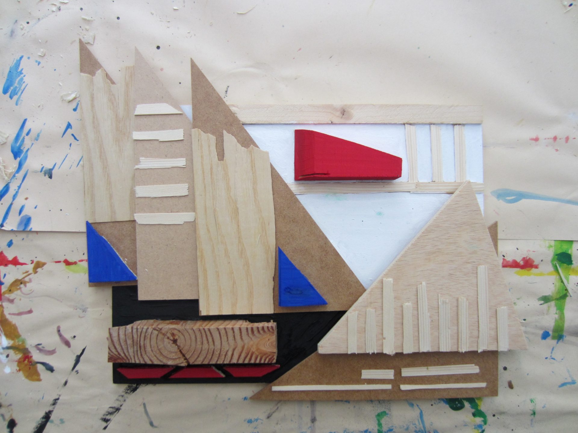 Ein abstraktes Mixed-Media-Kunstwerk aus hölzernen Dreiecken, Rechtecken und bemalten Formen in Rot, Blau, Schwarz und Weiß auf einer mit Farbe befleckten Oberfläche. Die Komposition ist geometrisch und vielschichtig.