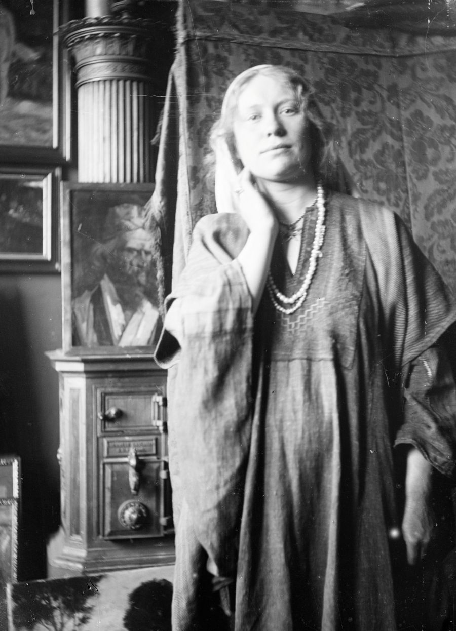 Ein Schwarz-Weiß-Foto im Vintage-Stil, das eine Frau in einem weiten Kleid und einer langen Halskette zeigt, die mit einer Hand am Hals posiert. Sie steht in einem Raum, der mit Gemälden, kunstvollen Möbeln und gemusterten Vorhängen geschmückt ist.