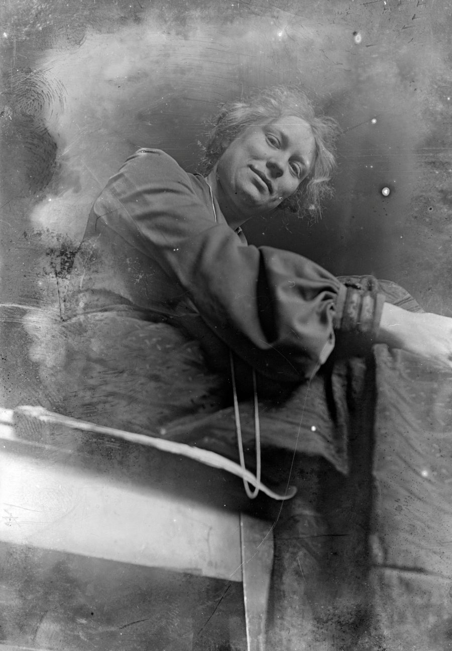 Ein schwarz-weißes Vintage-Foto einer Frau mit kurzen, gewellten Haaren und einem ruhigen Gesichtsausdruck, die sitzt und sich leicht nach vorne beugt; sie trägt ein langärmeliges Kleid und eine Halskette, vor einem verschwommenen, rauchigen Hintergrund.