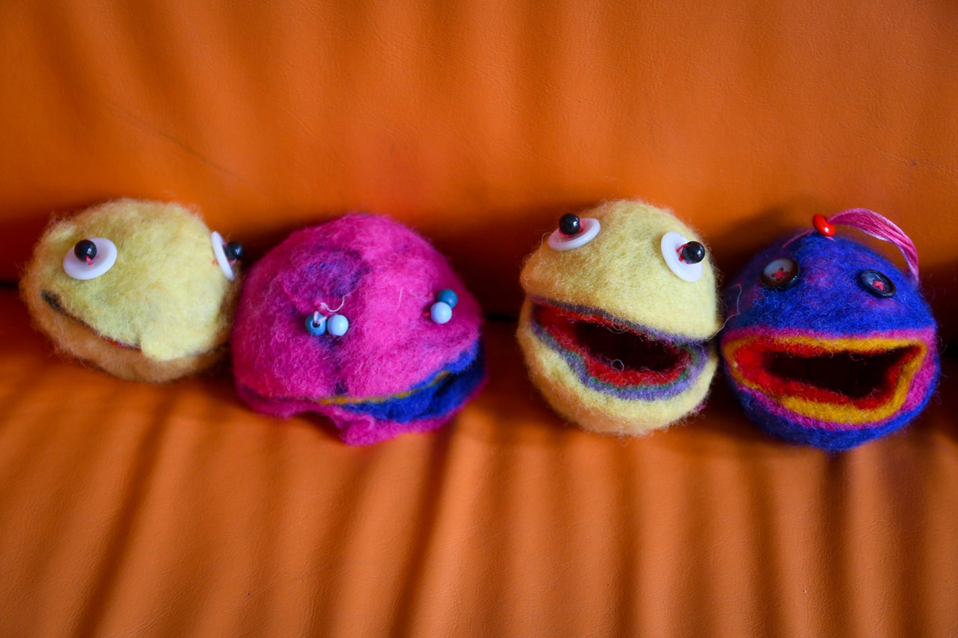 Vier bunte Filzpuppenköpfe mit Kulleraugen und offenen Mündern sind auf einer orangefarbenen Couch aufgereiht. Die Puppen sind gelb, rosa, gelb mit rot und blau mit roten Merkmalen.
