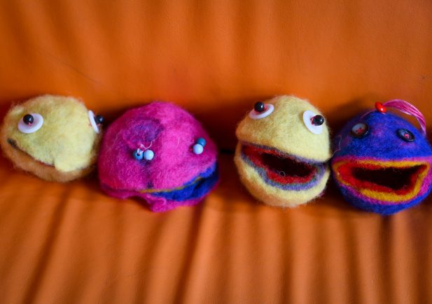 Vier bunte Filzpuppenköpfe mit Kulleraugen und offenen Mündern sind auf einer orangefarbenen Couch aufgereiht. Die Puppen sind gelb, rosa, gelb mit rot und blau mit roten Merkmalen.