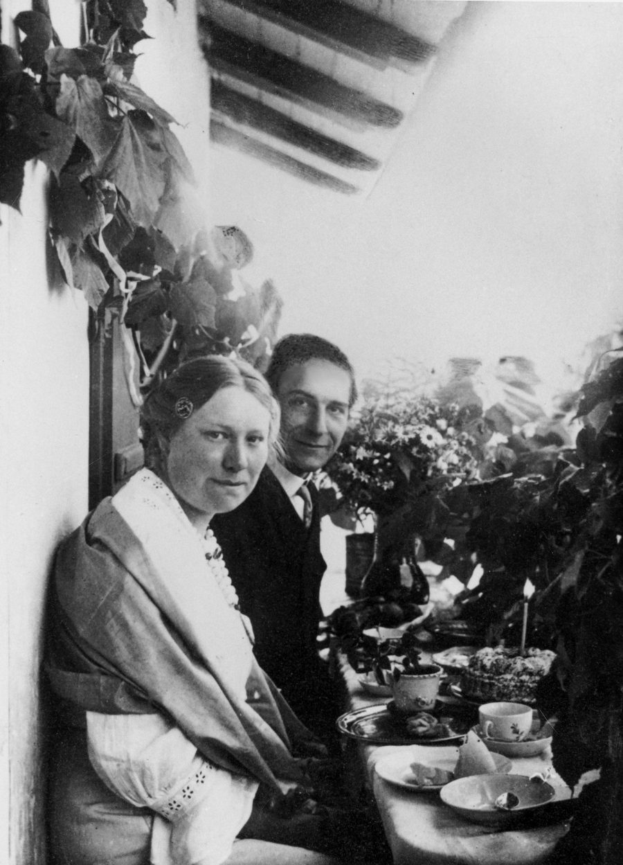 Ein Schwarz-Weiß-Foto einer Frau und eines Mannes, die an einem mit Tassen und Tellern gedeckten Tisch im Freien sitzen, umgeben von Pflanzen. Die Frau trägt einen Schal und blickt in die Kamera, während der Mann neben ihr lächelt.