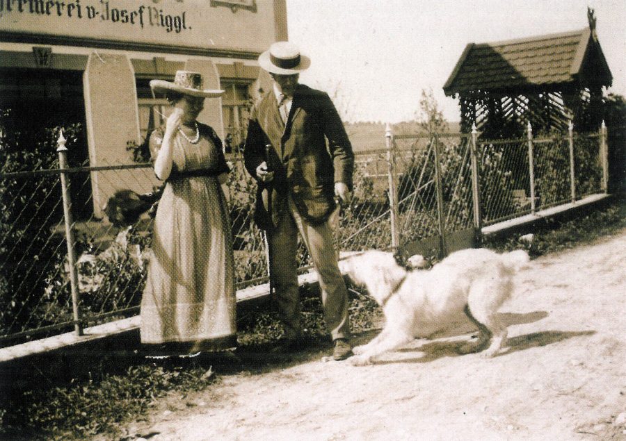 Eine Frau und ein Mann in Retro-Kleidung stehen auf einem Gehweg in der Nähe eines Zauns, während ein großer weißer Hund vor ihnen spielerisch an der Leine zerrt. Im Hintergrund sind ein Gebäude und Bäume zu sehen.