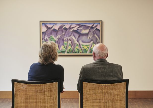 Zwei Personen sitzen nebeneinander auf Stühlen und blicken auf ein farbenfrohes Gemälde mit Eseln, das an einer schlichten Galeriewand hängt. Die Szene wird von hinten gezeigt und gibt den Rücken der beiden Personen wieder, während sie das Kunstwerk betrachten.