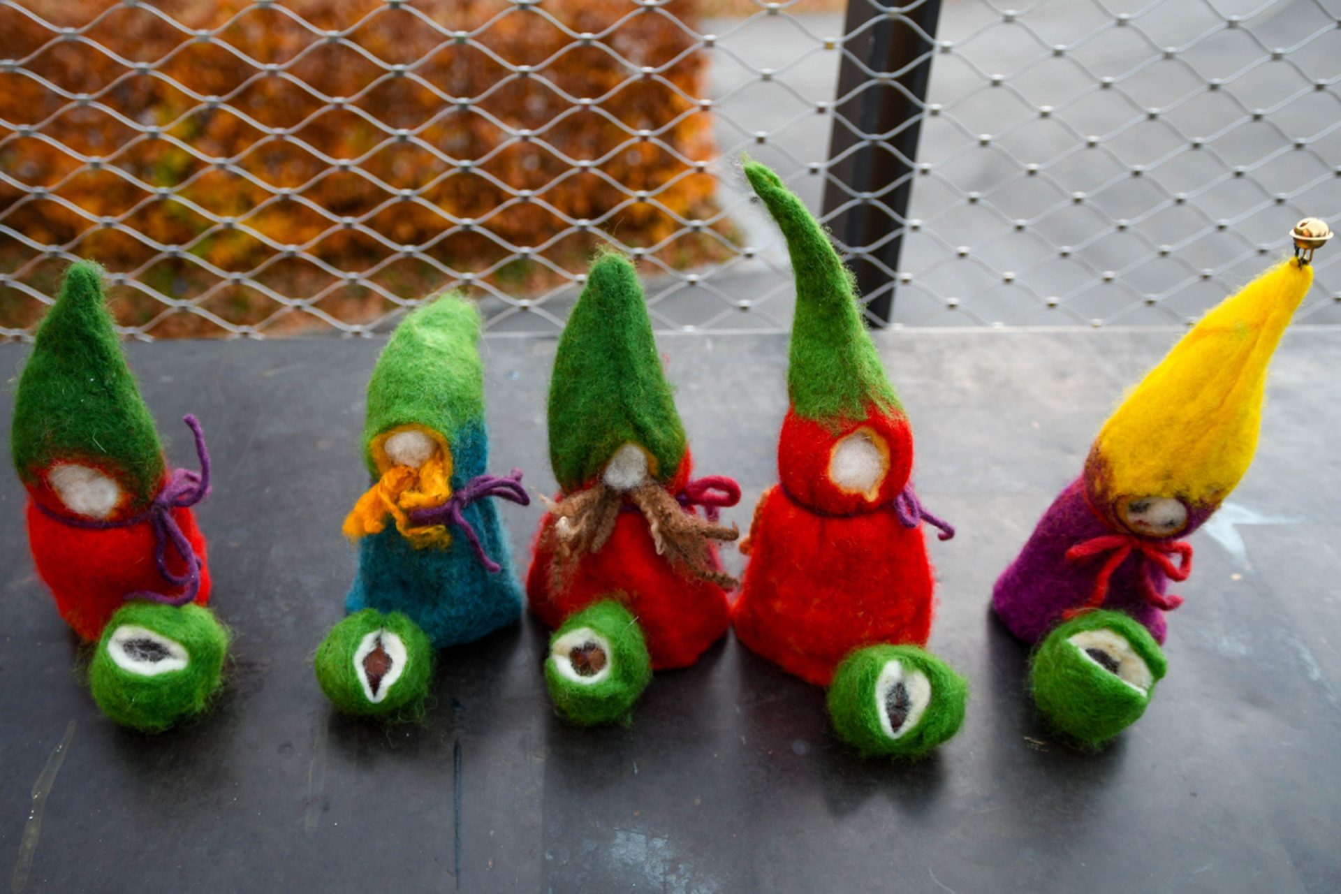 Fünf bunte Filz-Zwerge mit langen Hüten, jeweils zusammen mit einem kleinen grünen Filzkügelchen, stehen auf einem Tisch im Freien, im Hintergrund sind ein Drahtzaun und Herbstlaub zu sehen.