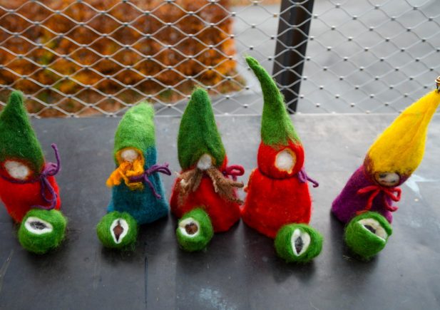 Fünf bunte Filz-Zwerge mit langen Hüten, jeweils zusammen mit einem kleinen grünen Filzkügelchen, stehen auf einem Tisch im Freien, im Hintergrund sind ein Drahtzaun und Herbstlaub zu sehen.