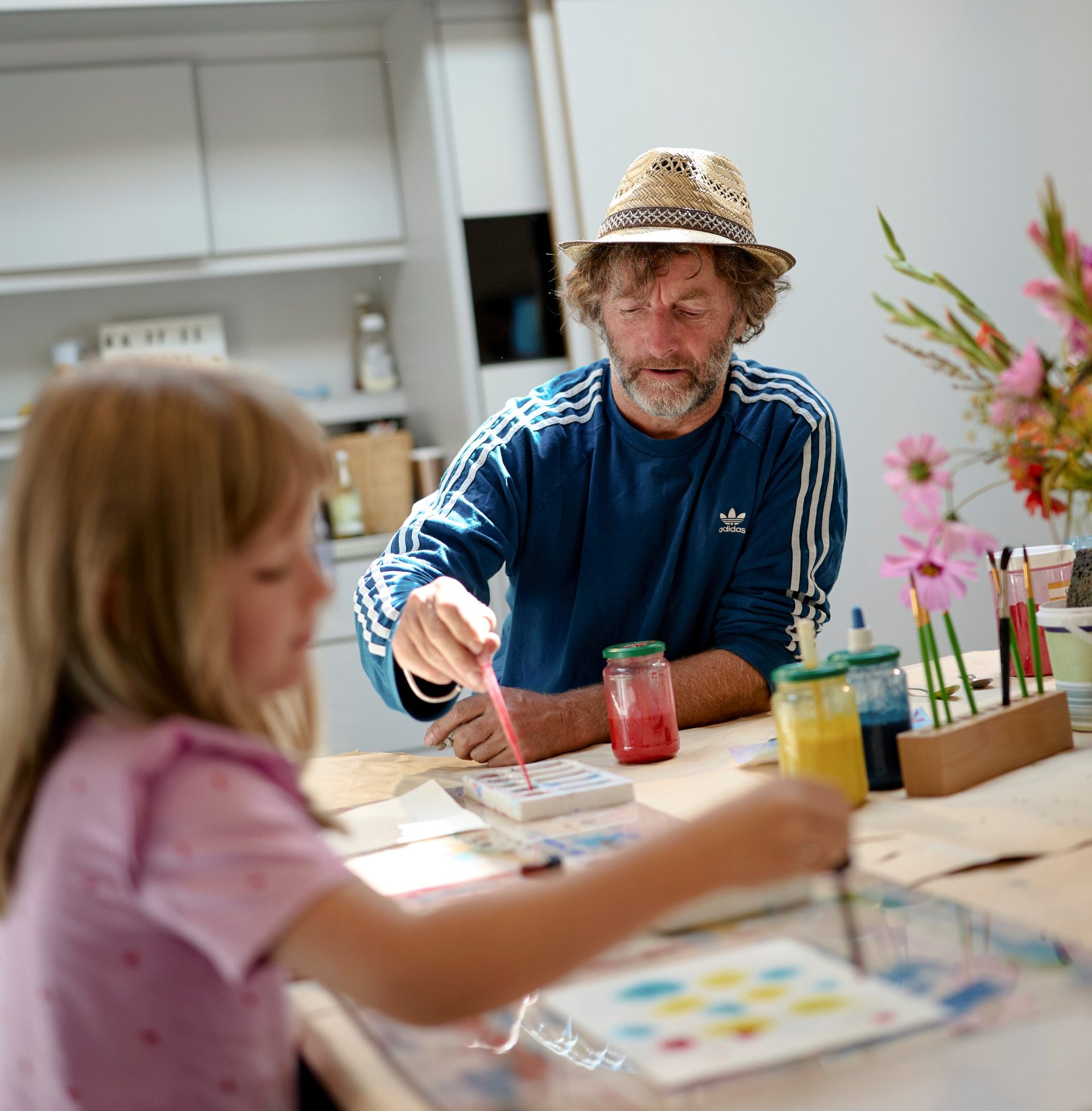 Ein Mann mit Strohhut und blauem Hemd malt zusammen mit einem kleinen Mädchen an einem Tisch. Auf dem Tisch liegen Malutensilien und bunte Blumen.