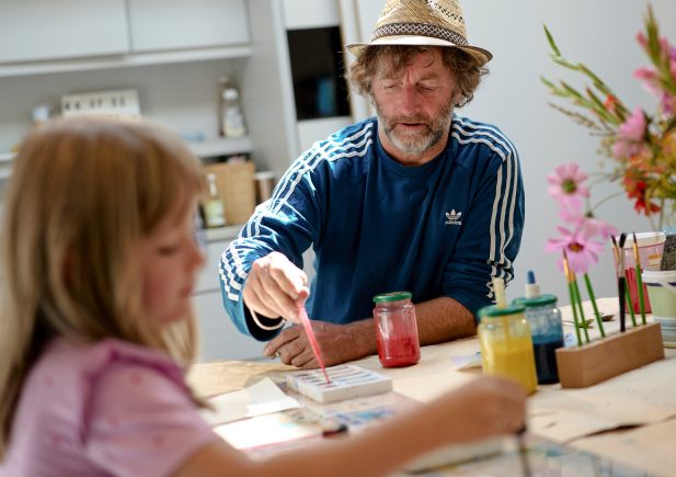 Ein Mann mit Strohhut und blauem Hemd malt zusammen mit einem kleinen Mädchen an einem Tisch. Auf dem Tisch liegen Malutensilien und bunte Blumen.