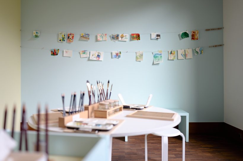 Ein runder Tisch mit Malutensilien im Vordergrund, im Hintergrund eine hellblaue Wand, an der Kinderzeichnungen an Schnüren befestigt sind.