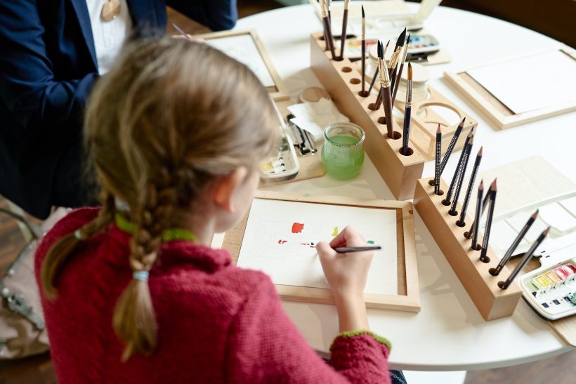 Ein junges Mädchen mit geflochtenen Zöpfen malt auf einer kleinen Leinwand an einem Tisch, auf dem Pinsel, Bleistifte und Aquarellfarben bereitliegen.