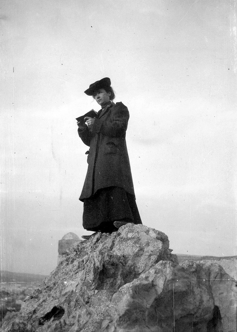Eine Frau in einem langen Mantel und mit Hut steht auf einem Felsvorsprung und blickt auf ein Buch hinab, das sie in den Händen hält.
