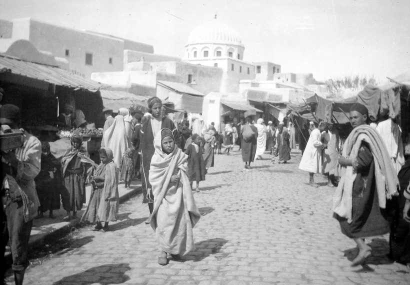 Menschen in traditioneller Kleidung gehen auf einer Kopfsteinpflasterstraße auf einem Markt entlang, im Hintergrund ist ein Kuppelgebäude zu sehen.